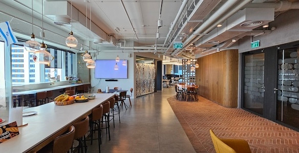 מטבח משרדי מודרני ופינת ישיבה עם בר, שולחנות עץ, מנורות תלייה, קערות פירות וחלונות גדולים המשקיפים על בנייני העיר — מושלם למשרד מרוהט במגדל סקיי ת״א.