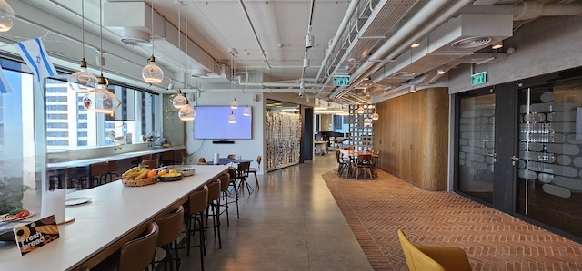 מטבח משרדי מודרני ופינת ישיבה עם בר, שולחנות עץ, מנורות תלייה, קערות פירות וחלונות גדולים המשקיפים על בנייני העיר — מושלם למשרד מרוהט במגדל סקיי ת״א.