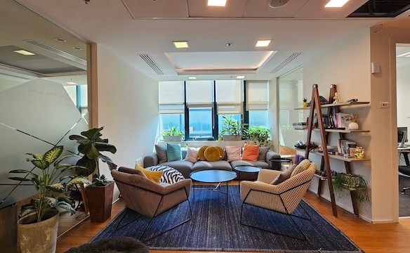 טרקלין משרדי מודרני בבית רובינשטיין עם ספה אפורה, שני כורסאות, שולחנות קפה עגולים, עציצים, שטיח כחול, מדף קיר וחלונות גדולים המכניסים אור טבעי — מושלם למשרד להשכרה.