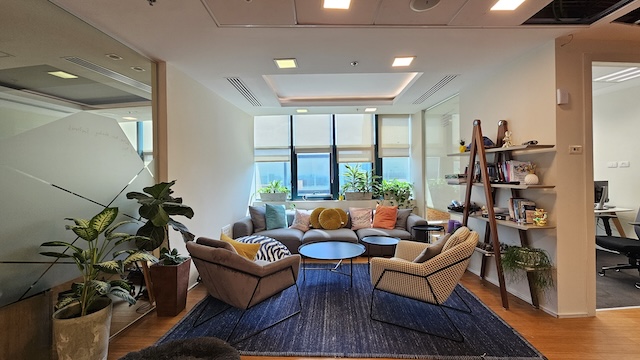 טרקלין משרדי מודרני בבית רובינשטיין עם ספה אפורה, שני כורסאות, שולחנות קפה עגולים, עציצים, שטיח כחול, מדף קיר וחלונות גדולים המכניסים אור טבעי — מושלם למשרד להשכרה.