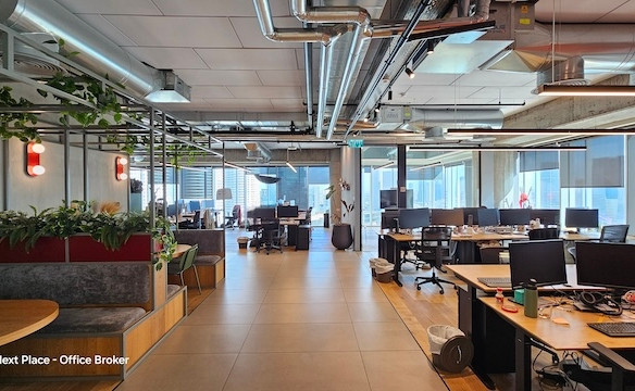 משרד מודרני בעל חלל פתוח עם שולחנות, כיסאות, מחשבים, תאורה תקרתית וצמחים פנימיים במגדל אקרו. החלל מואר ומסודר, עם חלונות גדולים ותעלות אוורור גלויות על התקרה. אידיאלי להשכרה בתל אביב.