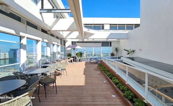 מרפסת גג חיצונית עם שולחנות, כיסאות, עציצים ומעקה זכוכית על גג מגדל אקרו, המשקיפה על נוף העיר תחת שמיים כחולים וצלולים.