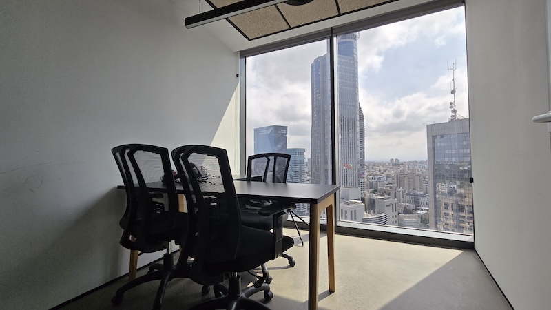 חדר ישיבות קטן ומרוהט עם שלושה כיסאות משרדיים ושולחן, עם חלון גדול המשקיף על מגדל ספיר, הבורסה ונוף העיר.