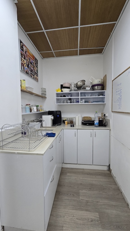 מטבח קטן במשרד להשכרה ברמת גן, מתחם הבורסה, עם ארונות לבנים, מכשירי חשמל על השיש, כלים על מדפים, מתלה כלים, לוח לבן על הקיר ותמונות המוצגות מעל מדף.