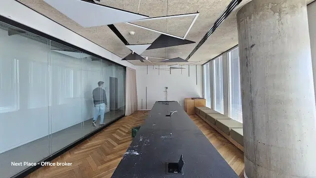 חדר ישיבות מודרני עם שולחן שחור ארוך, ספסלי ישיבה ליד החלונות, לוחות תקרה גיאומטריים, ואדם הולך ליד קיר זכוכית במשרד גדול להשכרה במגדל עזריאלי שרונה.