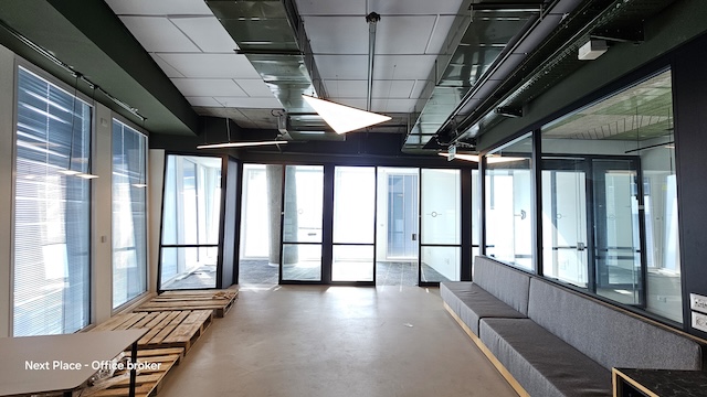 משרד גדול במגדל עזריאלי שרונה עם חלונות גדולים, דלתות זכוכית, ספסל ארוך אפור, תקרה חשופה ומשטחי עץ בפינה.