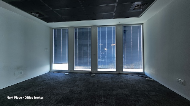 משרד מרווח וריק בשטח של 1200 מ"ר עם שטיח כהה, קירות לבנים וארבעה חלונות גבוהים המכוסים בתריסים אנכיים כחולים במגדל עזריאלי שרונה; אור טבעי מינימלי ממלא את המשרד הגדול הזה.