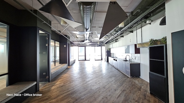 חלל משרדי מודרני בעל תכנון פתוח עם רצפות עץ, חלונות גדולים, תאורה תקרתית, צנרת חשופה ואזור מטבחון — אידיאלי עבור משרדים להשכרה או משרד גדול במגדל עזריאלי שרונה.