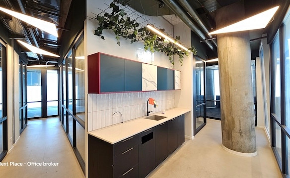 מטבחון משרדי מודרני עם ארונות מובנים, כיור ותאורה מתחת לארונות במשרד גדול של 1200 מ"ר במגדל עזריאלי שרונה. כולל עמוד בטון, משרדים עם קירות זכוכית, צמחייה על התקרה ותעלות אוורור חשופות.