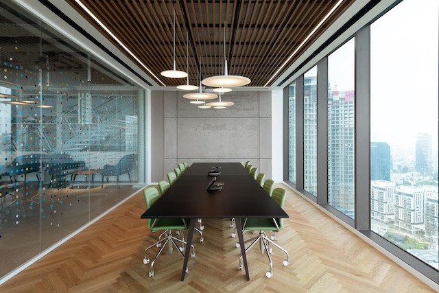 חדר ישיבות מודרני במגדלי הארבעה, הכולל שולחן שחור ארוך, כיסאות ירוקים, מנורות תלייה, לוחות תקרה מעץ וחלונות גדולים המשקיפים על נוף העיר המרהיב של שרונה. הזדמנות מושלמת למשרד למכירה.