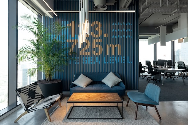 אזור ישיבה מודרני במשרד עם ספה, כיסאות ושולחן קפה ליד חלונות גדולים במגדלי הארבעה; על הקיר כתוב "1143.725 מ' מעל פני הים" — מושלם למי שמחפש משרד להשכרה קרוב לשרונה.