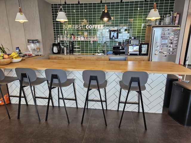 בר קפה מודרני עם דלפק עץ, חמישה כסאות בר אפורים, קיר אריחים ירוק ומכונות קפה — מושלם לכל משרד מעוצב או משרדים במגדלי הארבעה. על השלט כתוב "BUT FIRST, COFFEE" (אבל קודם, קפה). על המקרר יש מגנטים משעשעים.