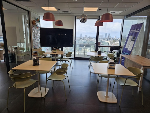 חדר מנוחה מעוצב ומודרני עם מספר שולחנות וכסאות, חלונות גדולים המשקיפים על מגדלי הארבעה, וטלוויזיה התלויה על הקיר.