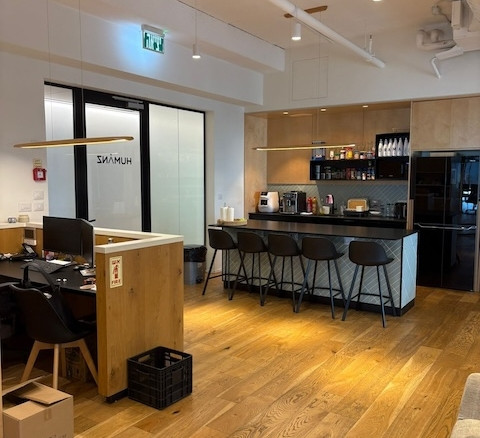 אזור מטבח משרדי מודרני עם דלפק, ארבעה כסאות בר, מכונות קפה, מדפים עם ציוד ותחנות עבודה סמוכות על רצפת עץ — אידיאלי עבור משרדים להשכרה במגדל רסיטל תל אביב.