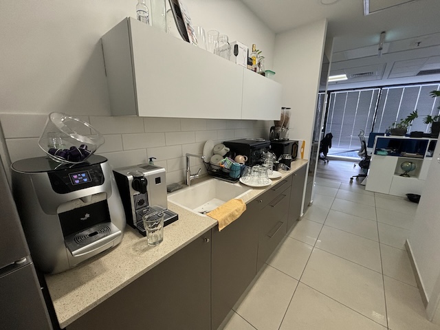 מטבחון משרדי עם מכונות קפה, כלים וכוסות על השיש משקיף על אזור משרדי סמוך עם שולחנות וחלונות גדולים — אידיאלי עבור מי שמחפש משרד מודרני להשכרה במגדלי אלון תל אביב.