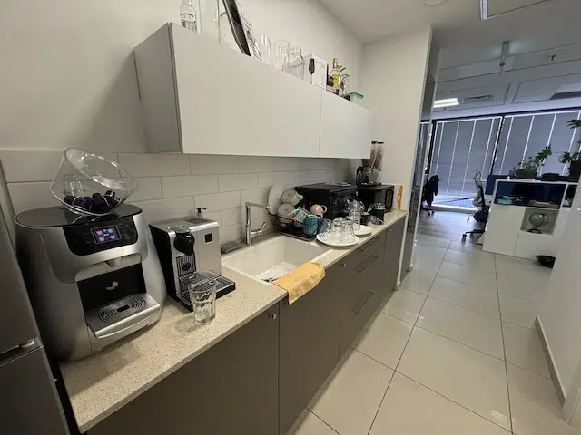מטבחון משרדי עם מכונות קפה, כלים וכוסות על השיש משקיף על אזור משרדי סמוך עם שולחנות וחלונות גדולים — אידיאלי עבור מי שמחפש משרד מודרני להשכרה במגדלי אלון תל אביב.