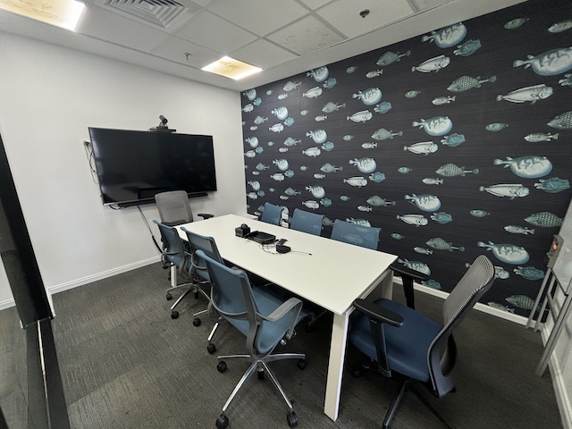 משרד קטן להשכרה במגדלי אלון, תל אביב, הכולל שולחן לבן ארוך, שבעה כיסאות משרדיים, טלוויזיה המותקנת על הקיר וקיר המעוטר בדוגמת דגים.