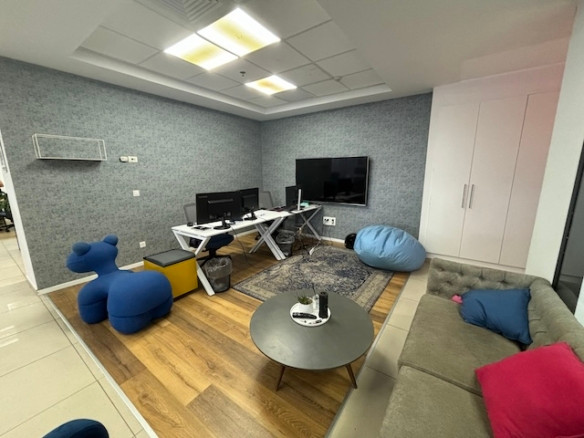 טרקלין משרדי מודרני במגדלי אלון, תל אביב, הכולל ספה אפורה, שולחן עגול, פוף כחול, כיסא בצורת חיה, שולחנות עם מסכים, טלוויזיה על הקיר וריצוף עץ ואריחים מסוגנן — משרד להשכרה אידיאלי.