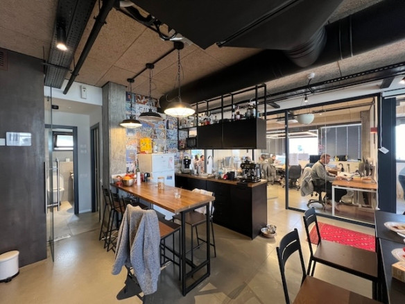 אזור מטבח משרדי מודרני עם שולחנות עץ, כיסאות ומכשירים חשמליים בקדמת התמונה; עובדים העובדים ליד שולחנות במשרד מרוהט בקירות זכוכית בשטח של 200 מ"ר ברוטשילד נראים ברקע.