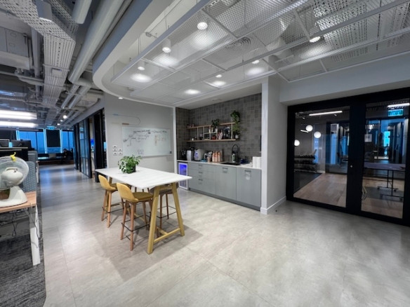 אזור מטבח משרדי מודרני עם שולחן לבן, כיסאות צהובים, מכונות קפה וארונות במשרד מרוהט; דלתות זכוכית חושפות חלל עבודה פתוח במגדלי סקיי, תל אביב.