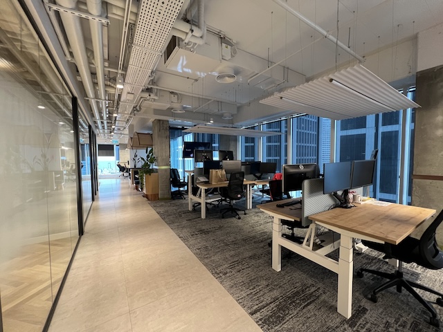 משרד מרוהט מודרני בעל חלל פתוח עם שולחנות ריקים, מחשבים וכסאות. חלונות גדולים משתרעים לאורך הצד הימני ומאפשרים כניסת אור טבעי. המשרד ממוקם במגדלי סקיי ת״א, וקיר זכוכית משתרע לאורך המסדרון השמאלי.