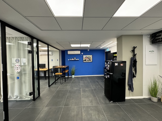 משרד מרוהט ומודרני ברחוב המסגר בתל אביב, עם קיר כחול בולט, שולחן גבוה עם שרפרפים, מקרר שחור, מתלה מעילים ועציצים על רצפת אריחים אפורים.