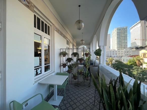 מרפסת מקורה ברוטשילד תל אביב עם כיסאות ירוקים, שולחנות לבנים, עציצים ונוף מקושת של בנייני העיר — מושלמת למשרד עם מרפסת או למשרד להשכרה.
