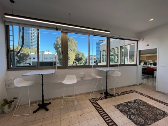 אזור ישיבה מודרני במשרד עם כיסאות לבנים ושולחנות גבוהים ליד חלונות גדולים, רצפת אריחים ונוף של בניינים ועצים בחוץ, הממוקם בבניין עצמאי ברחוב רוטשילד בתל אביב.