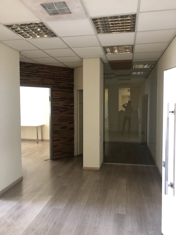 מסדרון בבית הקרן, עם ריצוף עץ בהיר, קירות לבנים, קיר עץ דקורטיבי, דלתות זכוכית ותאורת תקרה פלורוסנטית. אדם נראה בהשתקפות — משרד להשכרה במתחם הבורסה.