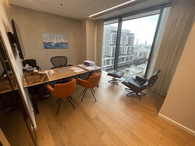 משרד להשכרה בשרונה, במגדלי הארבעה: עיצוב מודרני עם שולחן עץ, כיסאות כתומים, כורסת לאונג' ליד חלון גדול ונוף עירוני. על השולחן ניירת ומחשב; על הקיר תלויה יצירת אומנות של מפרשית.