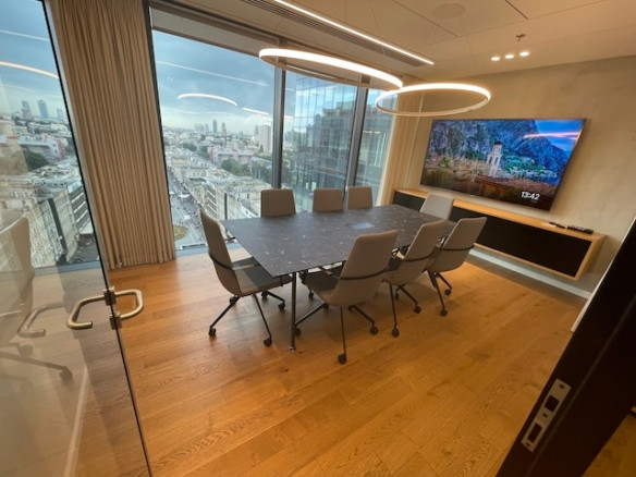 חדר ישיבות מודרני עם שולחן גדול, שמונה כיסאות, רצפת עץ, טלוויזיה המותקנת על הקיר וחלונות מהרצפה לתקרה המשקיפים על מגדלי הארבעה ושרונה — משרד להשכרה מושלם בלב העיר.