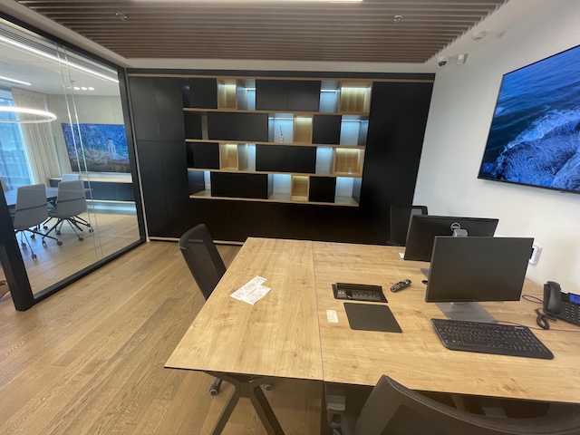 משרד להשכרה מודרני עם שני שולחנות, מסכים, מקלדת וטלפון. מדפים ותאורה מובנית מקשטים את הקיר האחורי; חדר ישיבות אלגנטי נראה מבעד לקירות הזכוכית במגדלי הארבעה, שרונה.