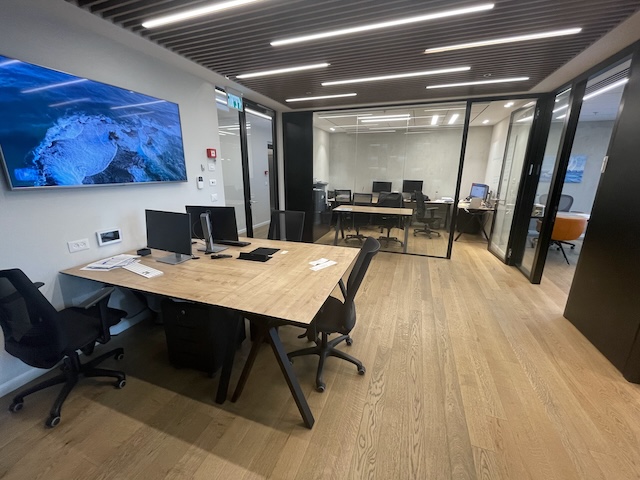 משרד מודרני עם רצפות עץ, מספר שולחנות וכסאות, מסך גדול המותקן על הקיר, וחדרי ישיבות עם קירות זכוכית ברקע במגדלי הארבעה שרונה – משרד להשכרה.