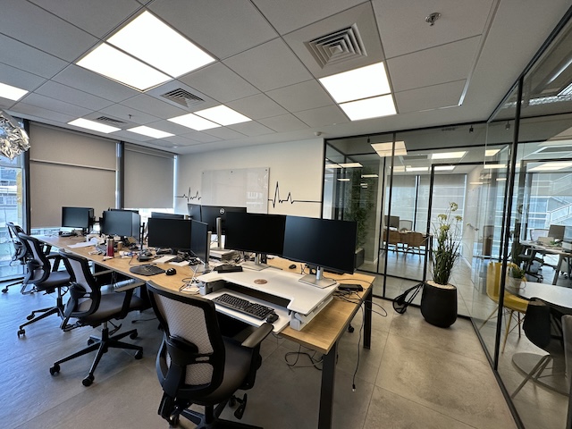 שטח משרדי מודרני להשכרה במגדלי הארבעה, תל אביב, הכולל שולחנות עבודה, מסכי מחשב, כיסאות משרדיים, חדר ישיבות עם קירות זכוכית ועציצים. מואר היטב בתאורת תקרה ובאור טבעי מהחלונות.