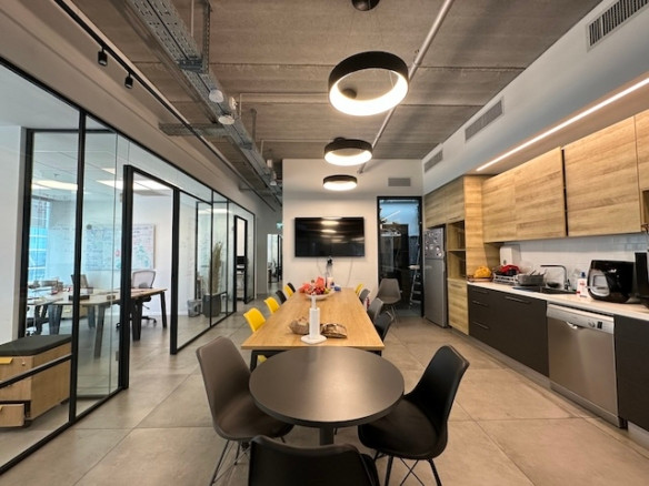 מטבח ומטבחון מודרניים במגדלי הארבעה בתל אביב, הכוללים שולחן עץ ארוך, כיסאות שחורים וצהובים, מטבחון, שולחן עגול וחדרי ישיבות עם קירות זכוכית – מושלם עבור מחפשי משרד להשכרה.