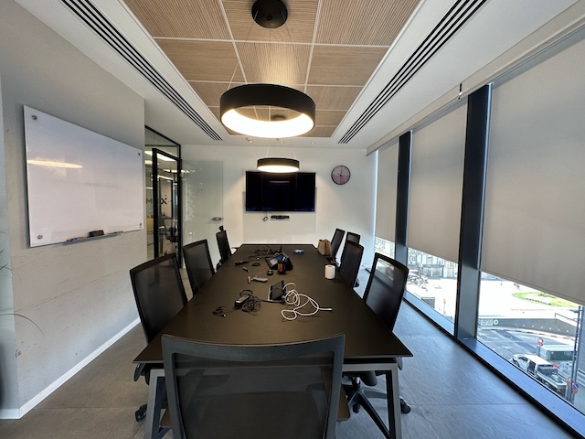 חדר ישיבות מודרני במגדלי הארבעה, תל אביב, הכולל שולחן ארוך, מספר כיסאות, לוח לבן, מסכים המותקנים על הקיר, שעון וחלונות גדולים עם תריסים. כבלים ומכשירים מונחים על השולחן — משרד להשכרה אידיאלי.