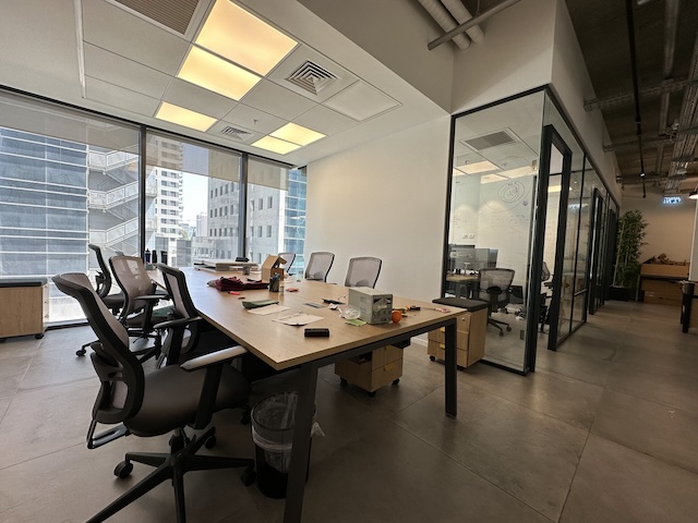חדר ישיבות משרדי מודרני עם שולחן גדול, מספר כיסאות, ציוד משרדי מפוזר וקירות זכוכית המשקיפים על מגדלי הארבעה בתל אביב — משרד להשכרה אידיאלי עם נוף לעיר מבעד לחלונות גדולים.