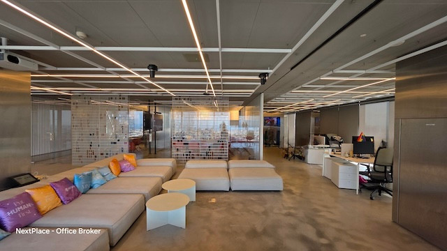 משרד להשכרה בתל אביב במגדלי הארבעה, עם ספות אפורות, כריות צבעוניות, שולחנות עגולים לבנים, מחיצות זכוכית, עמדות עבודה ותאורה בתקרה בסידור קווי.