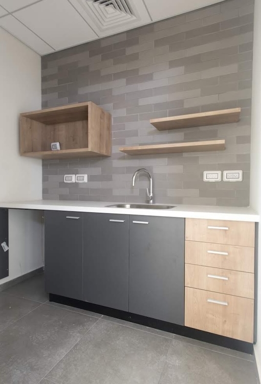 מטבחון מודרני עם ארונות אפורים ועץ, משטח עבודה לבן, כיור ומדפי עץ על קיר אריחים אפורים — מושלם למשרד להשכרה במגדל WE ת״א.