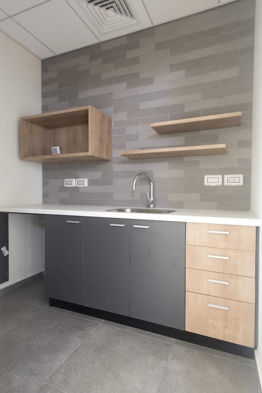מטבחון מודרני עם ארונות אפורים ועץ, משטח עבודה לבן, כיור ומדפי עץ על קיר אריחים אפורים — מושלם למשרד להשכרה במגדל WE ת״א.