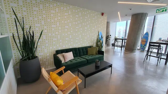 טרקלין משרדי מודרני עם ספה ירוקה, כיסא עץ, שולחן קפה שחור, עציץ גדול וקיר מעוטר. חלונות גבוהים משקיפים על העיר – מושלם עבור משרד מרוהט או משרדים להשכרה במשרד במגדל תוהא.