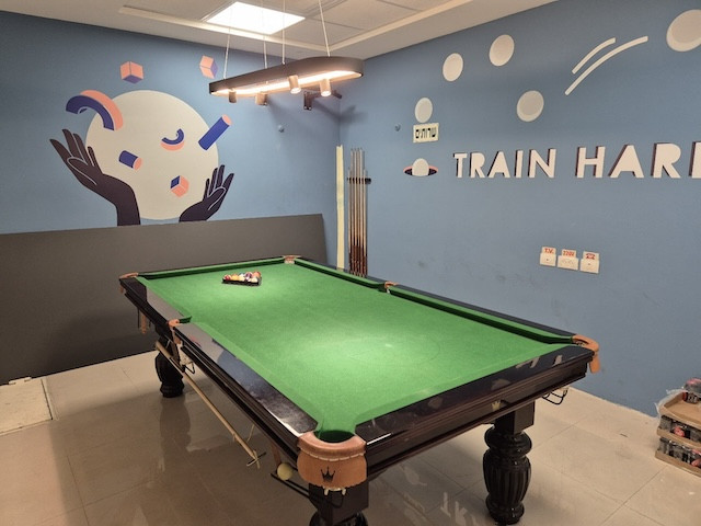 שולחן ביליארד עם לבד ירוק מוצב במשרד להשכרה ברמת גן, עם אמנות קיר וכתובת "TRAIN HARD" על הקיר. מקלות הביליארד מאוחסנים בצורה מסודרת בפינה.