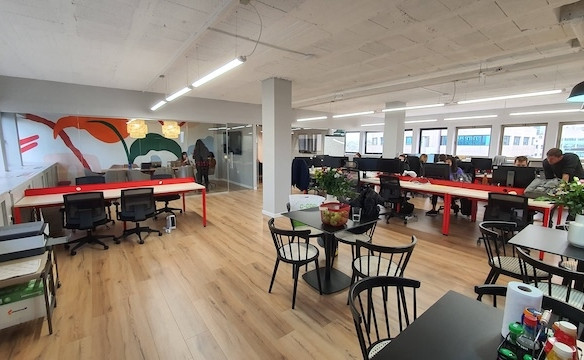 משרד להשכרה מודרני בעל חלל פתוח ברחוב החשמונאים, עם שולחנות, כיסאות, מחשבים ואנשים עובדים; צמחים וציוד על השולחנות; חלונות גדולים ומרפסת המספקים אור טבעי בשפע.