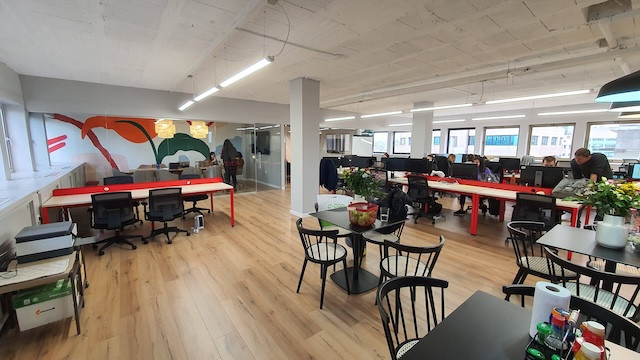 משרד להשכרה מודרני בעל חלל פתוח ברחוב החשמונאים, עם שולחנות, כיסאות, מחשבים ואנשים עובדים; צמחים וציוד על השולחנות; חלונות גדולים ומרפסת המספקים אור טבעי בשפע.