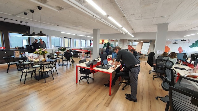 משרד פתוח עם אנשים העובדים ליד שולחנות, חלקם משתפים פעולה ואחרים מתרכזים במחשבים; עיצוב מודרני עם חלונות גדולים ורצפת עץ — משרד להשכרה אידיאלי בתל אביב.