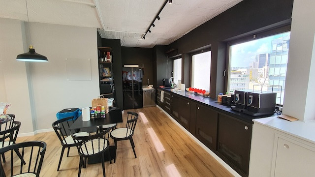 מטבח משרדי מודרני עם ארונות שחורים ומסוגננים, שולחן אוכל עם כיסאות, מכשירי חשמל שונים על השיש וחלונות גדולים המשקיפים על הבניינים ברחוב החשמונאים. מושלם למשרד להשכרה עם תאורה טבעית בשפע.