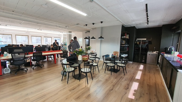 חלל משרדי פתוח עם שולחנות בצד שמאל, מטבח ופינת אוכל במרכז, ואנשים עובדים — הכל במשרד להשכרה ברחוב החשמונאים, חלקם נהנים מאוויר צח במרפסת.