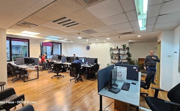 משרד להשכרה מודרני בעל חלל פתוח עם שולחנות עבודה רבים, עובדים העובדים מול מחשבים, ואדם אחד ליד מדף; כולל ריצוף עץ, קירות לבנים ואווירה מוארת.