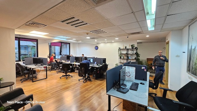משרד להשכרה מודרני בעל חלל פתוח עם שולחנות עבודה רבים, עובדים העובדים מול מחשבים, ואדם אחד ליד מדף; כולל ריצוף עץ, קירות לבנים ואווירה מוארת.