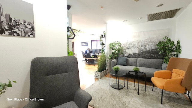 טרקלין משרדי מודרני עם כיסאות אפורים וכתומים, ספה, שולחנות קטנים וצמחים ירוקים. תמונות בשחור-לבן של נוף עירוני מקשטות את הקירות; ברקע נראים חלל עבודה פתוח ומרפסת. משרד להשכרה אידיאלי בבית אגיש רבד.